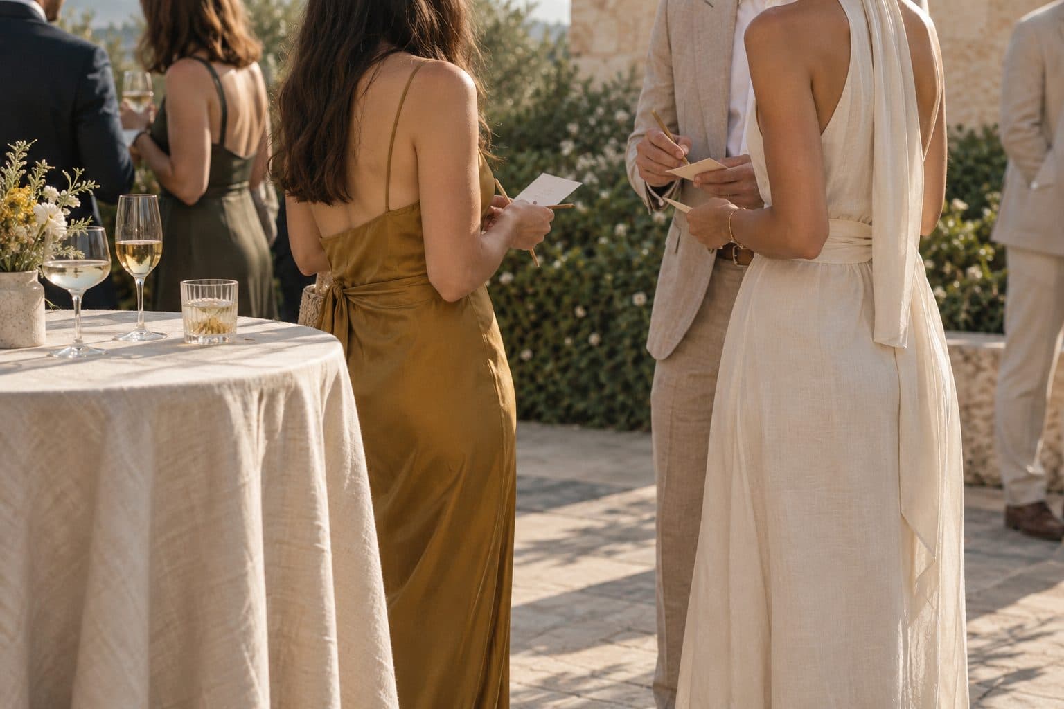 Invitados de pie en un cóctel al aire libre, vistos de espaldas, con cartones de papel y lápiz buscándose entre ellos: dinámicas para bodas